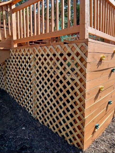 Wooden deck with lattice skirting and vertical railing, shown beside a dark mulch bed.