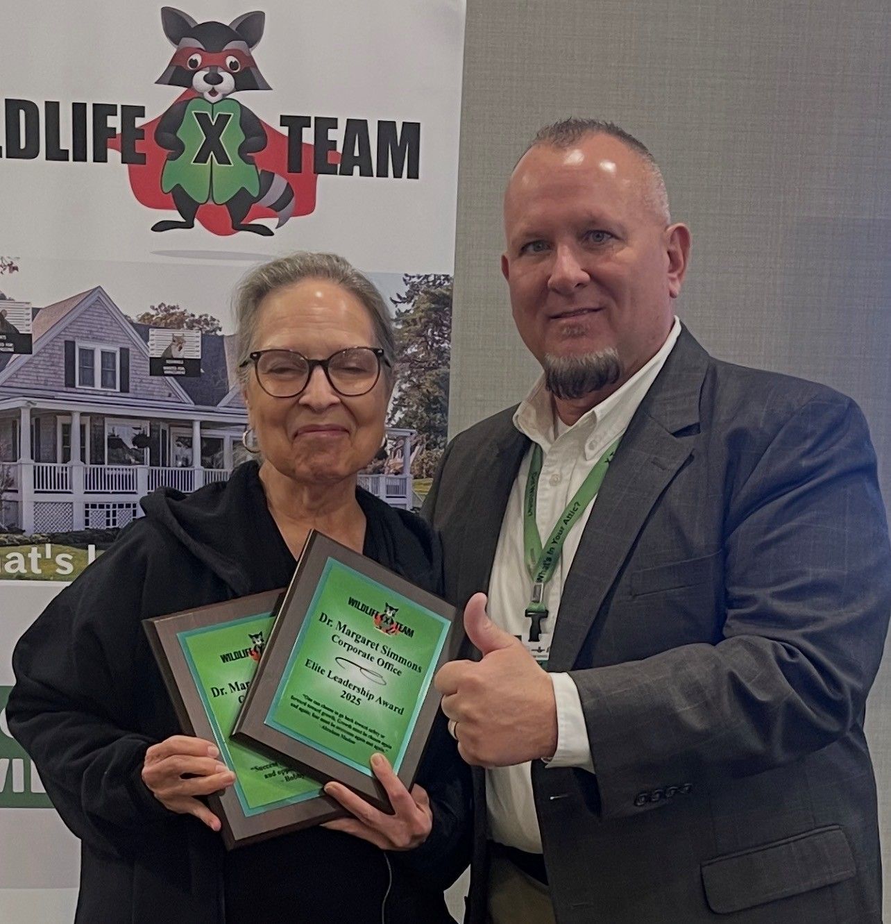 Two people holding framed awards in front of a Life Team banner, one giving a thumbs-up