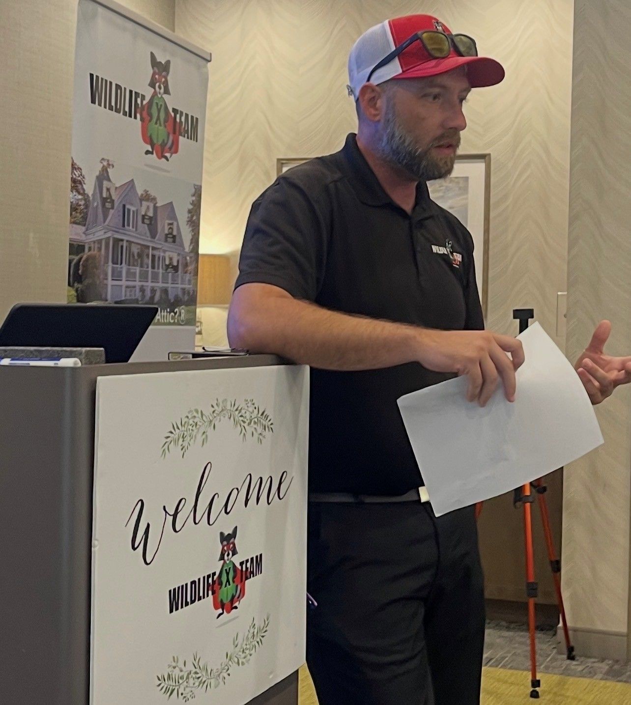 Man speaking at a welcome podium, holding papers in a hotel event room.