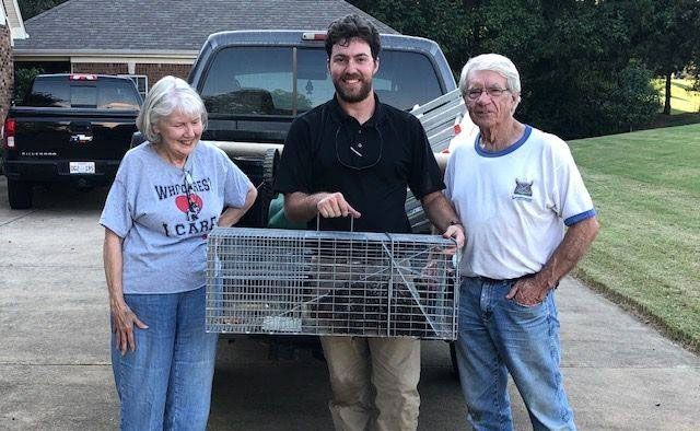 Three people holding a metal live trap outdoors in a driveway, with a pickup truck behind them