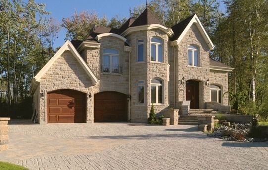 Stone house with brown garage doors and a paved driveway, surrounded by trees.