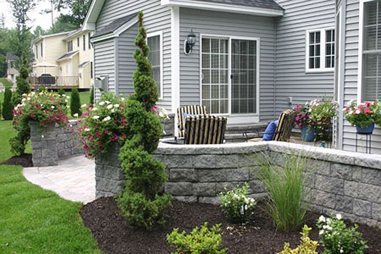 Stone patio with tiered planters and gray siding on a house. Green grass and shrubs.