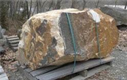 Large, multi-colored boulder strapped to a wooden pallet. Outdoor setting, trees in background.