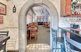 View into Italian restaurant dining area through stone archway. Tables, chairs, bar stools visible.