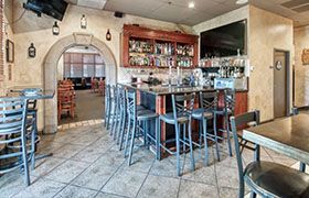 Bar interior with bar stools, shelves of alcohol, and an archway to another room.