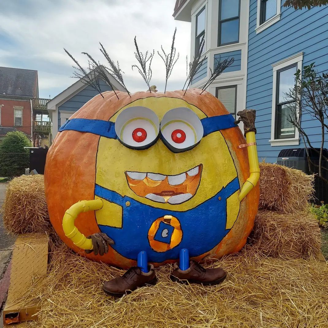 A giant pumpkin carved and painted to resemble a Minion sits on hay bales in front of a blue house.