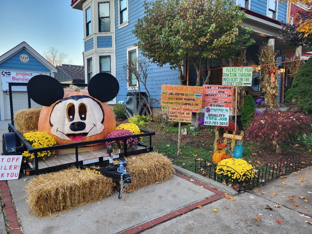A large Mickey Mouse-shaped pumpkin decoration sits on a trailer with hay bales and mums in front of a blue house.