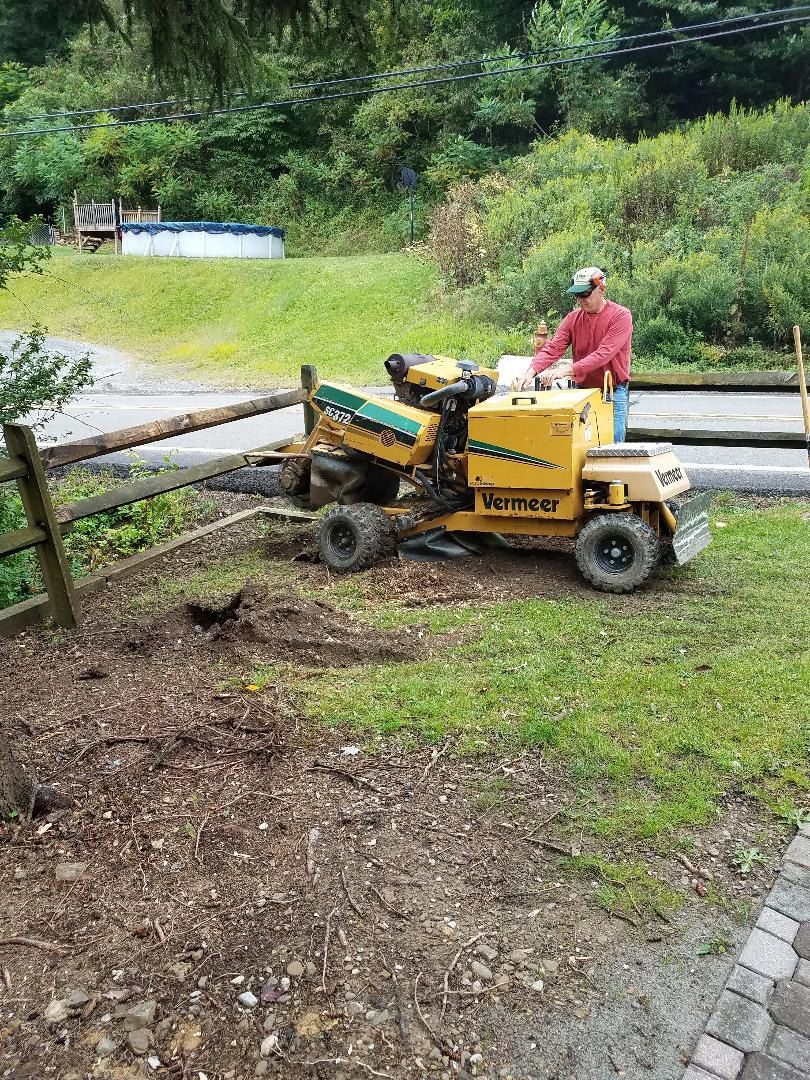 A person in a red shirt operates a yellow Vermeer stump grinder next to a wooden fence on a grassy hillside.