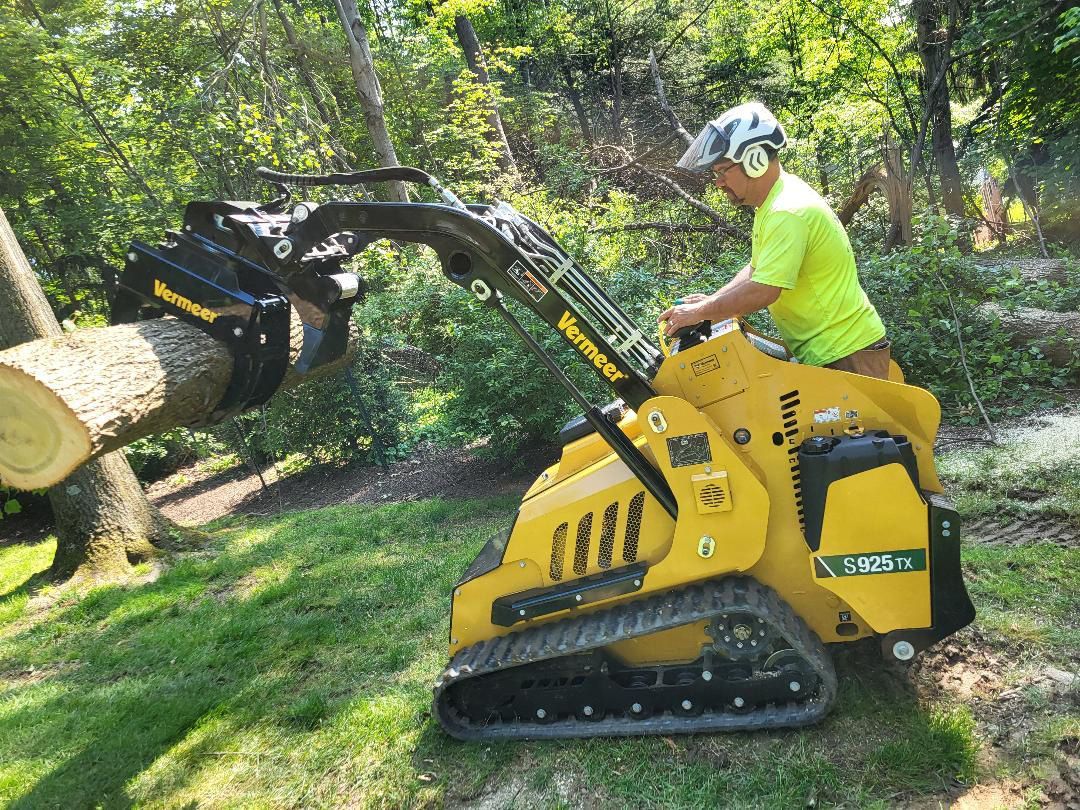 A person in a bright yellow shirt operates a yellow Vermeer tracked skid steer, using a grapple to lift a cut log.