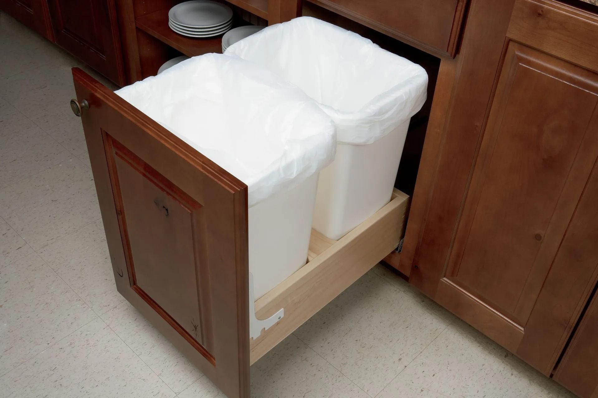 Cabinet door open revealing pull-out trash bins with white bags. Wooden cabinet.