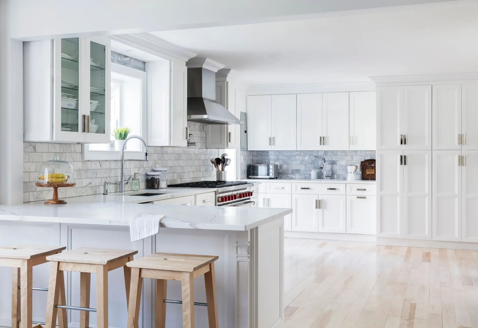 White kitchen with marble countertops, wood stools, stainless steel appliances, and a light wood floor.