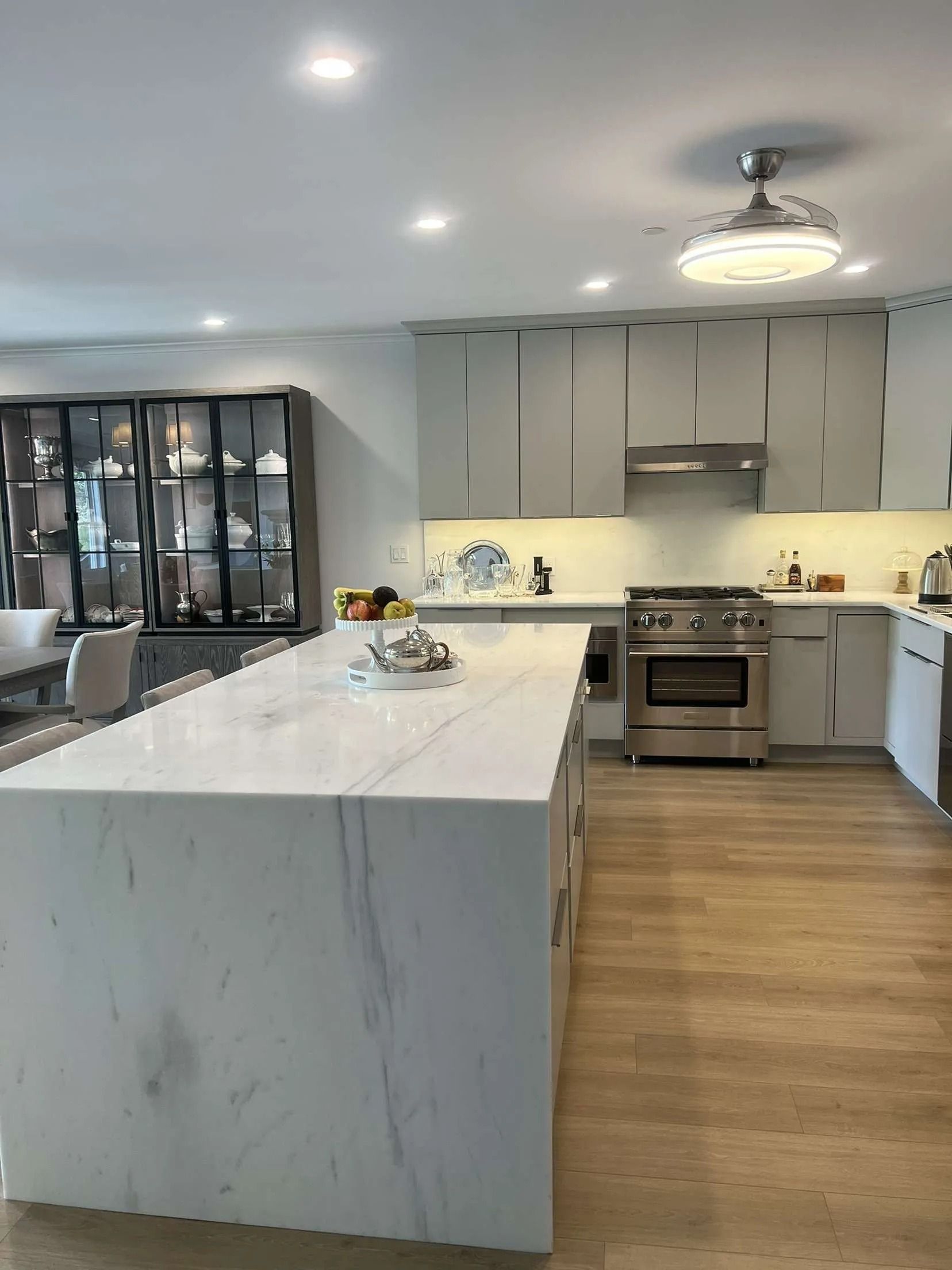 Modern kitchen with large marble island and light gray cabinets. Stainless steel appliances and wood flooring.
