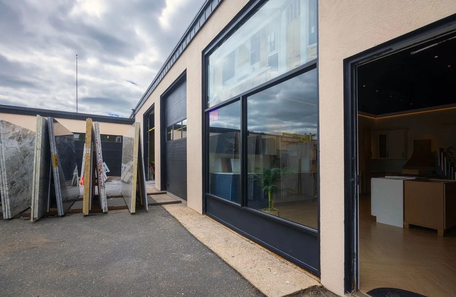 Exterior view of a stone showroom with large windows and open door. Slabs of stone stand outside. Cloudy sky.