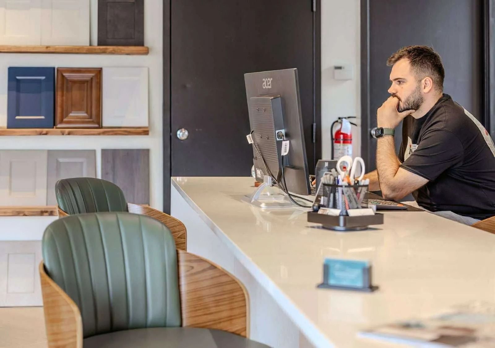 Man at computer in office, with samples on shelf. Thinking with hand on face.