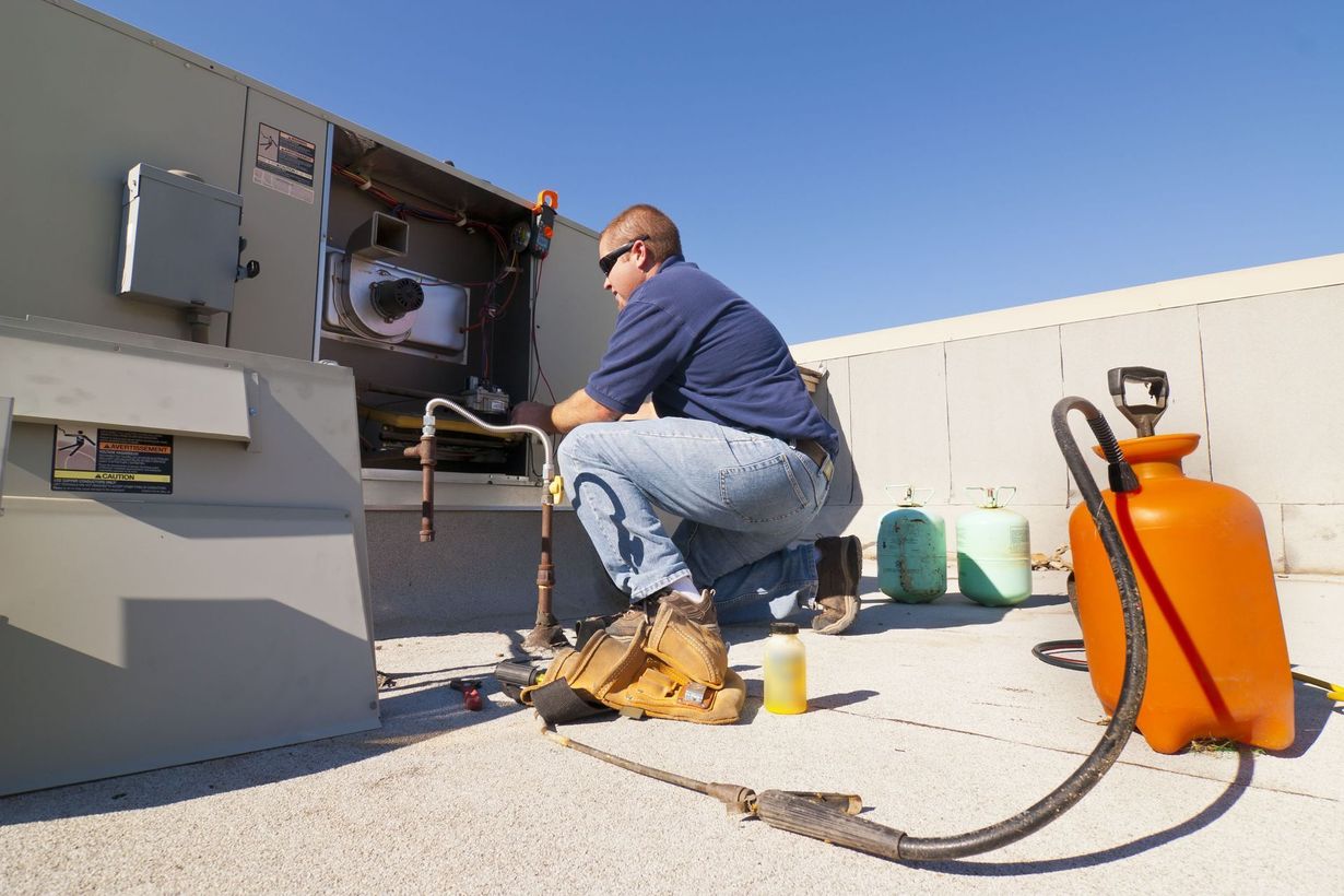 HVAC technician kneels, repairing an air conditioning unit on a rooftop, with tools and a container.