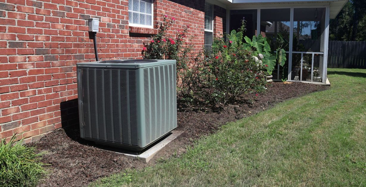 Air conditioning unit next to a brick wall and landscaping with mulch and plants.