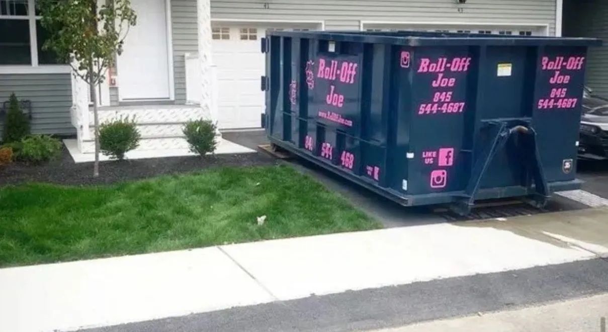 A large dark blue Roll-Off dumpster parked on the concrete driveway of a suburban house.