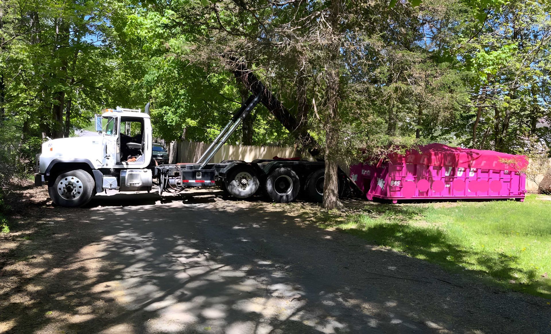 White truck with a pink dumpster in a wooded area, parked on a gravel driveway.