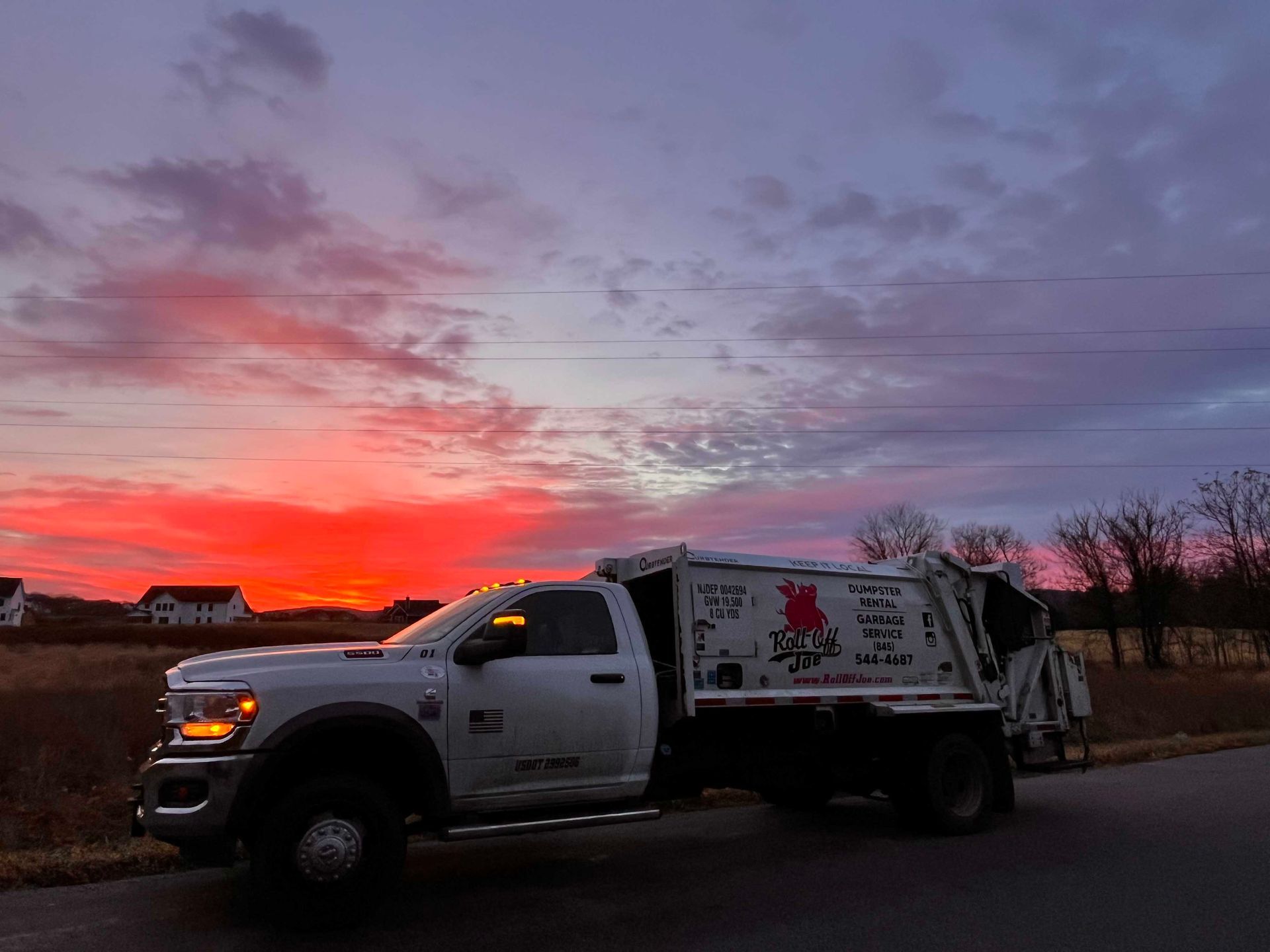 White garbage truck parked on road at sunset with red and orange sky.