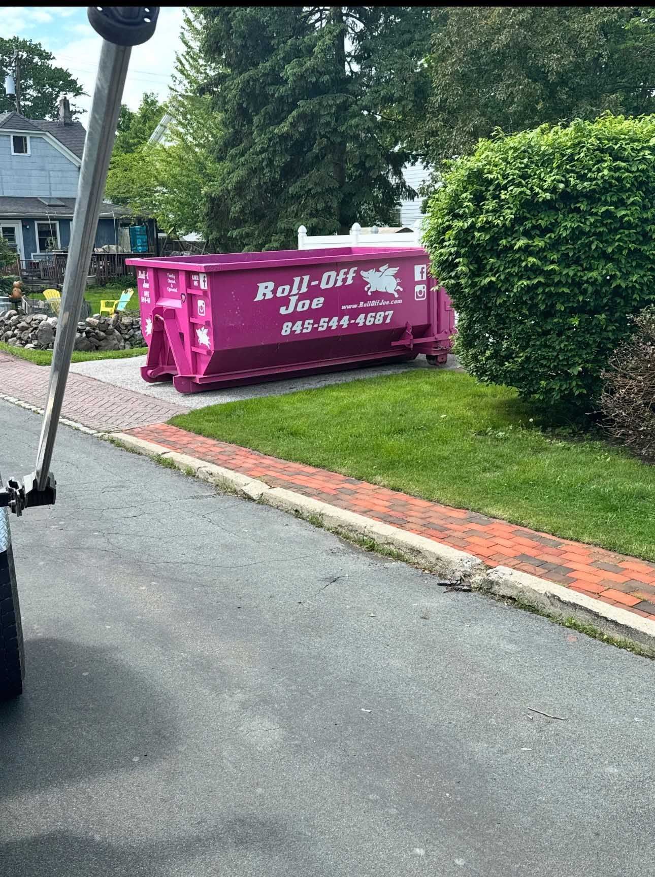 A bright pink Roll-Off Joe dumpster sits on a driveway next to a grassy area and brick border.