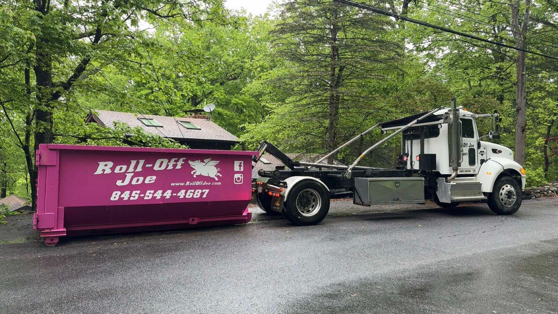 A pink Roll-Off Joe dumpster next to a white truck on a wet road, with green trees in the background.
