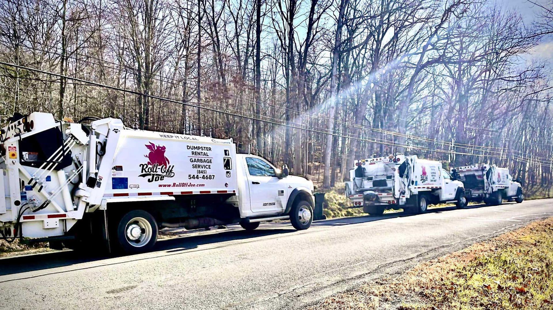 Three white garbage trucks parked on a road beside a wooded area. The first truck is closer, with detailed logos.