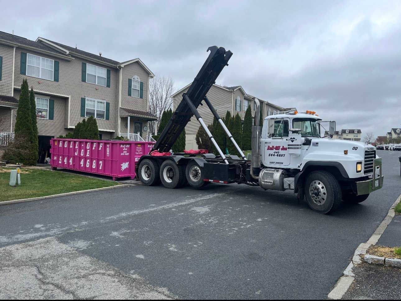 Pink dumpster being lifted by a white truck in front of a townhome on a cloudy day.
