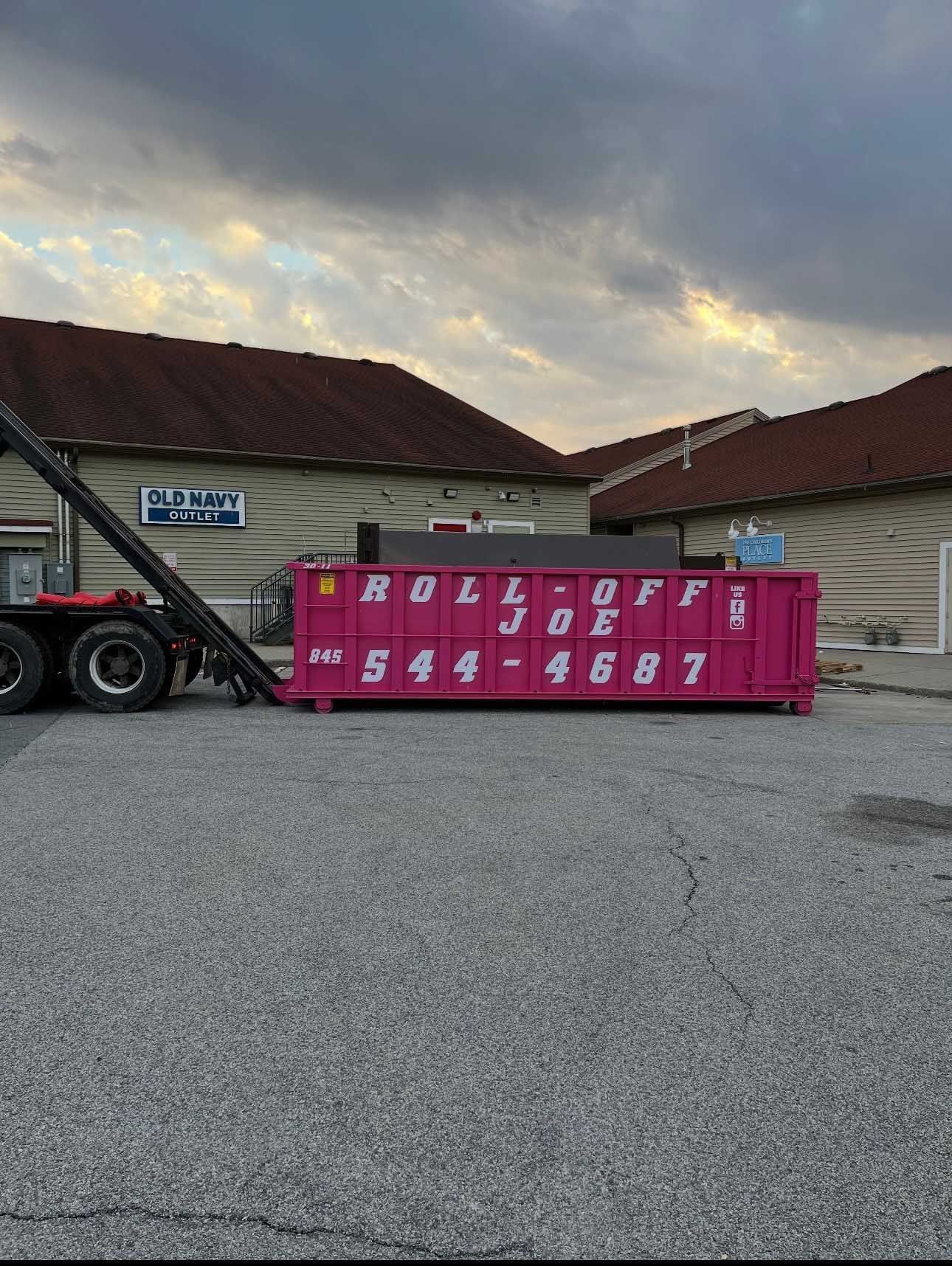 Pink dumpster being lifted by a truck in front of a building. 