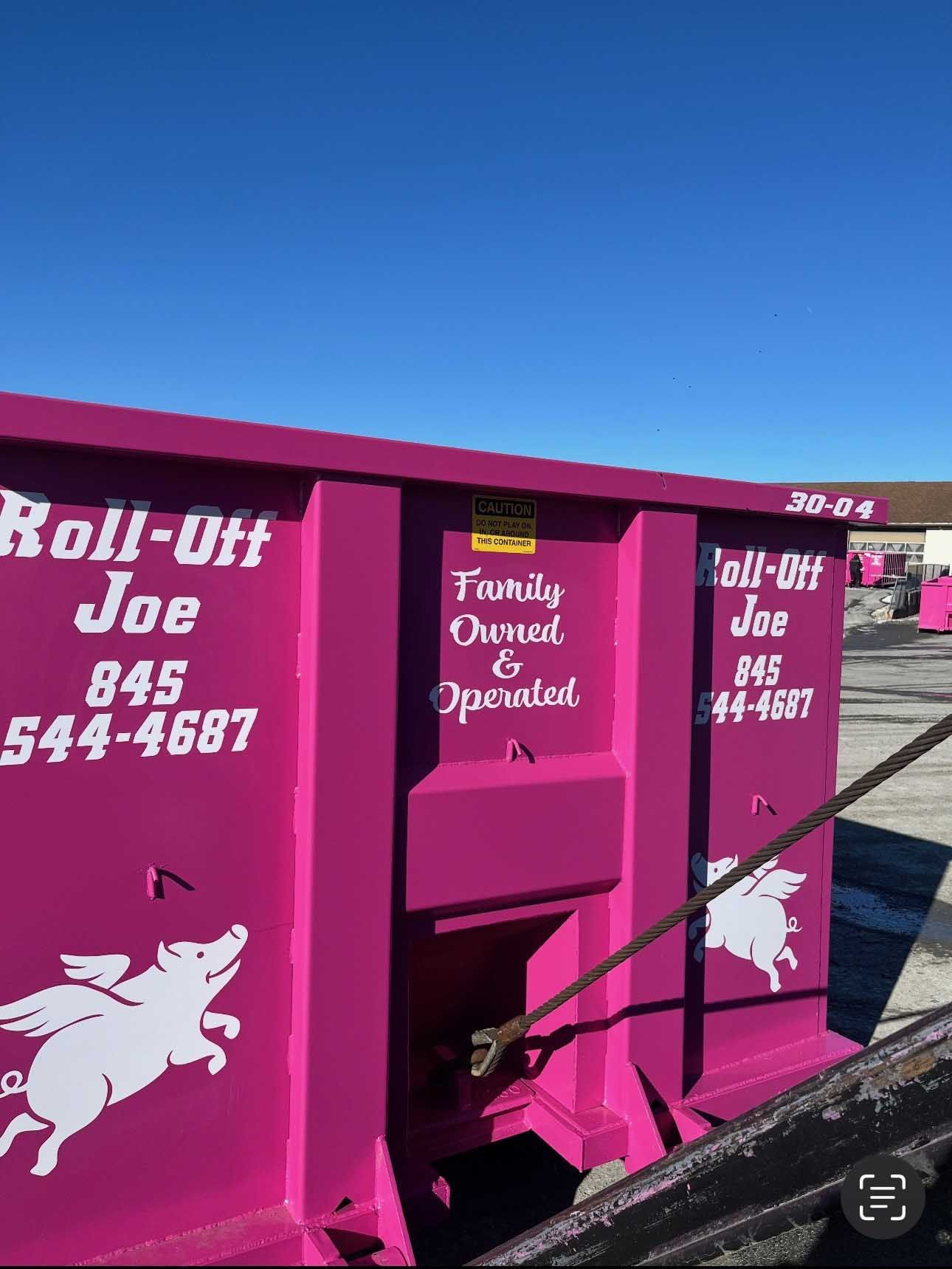 Hot pink Roll-Off Joe dumpster with white text, pig logo, and phone number; outdoors on a sunny day.