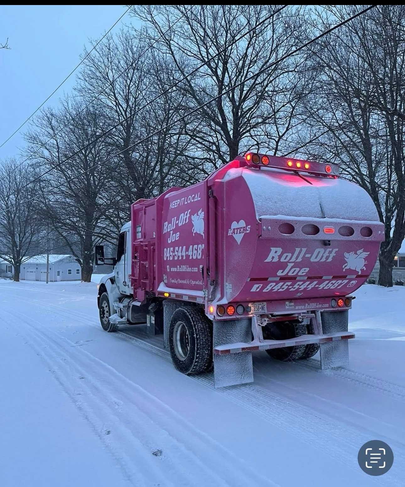 Pink garbage truck driving on a snow-covered road. The truck has the words 