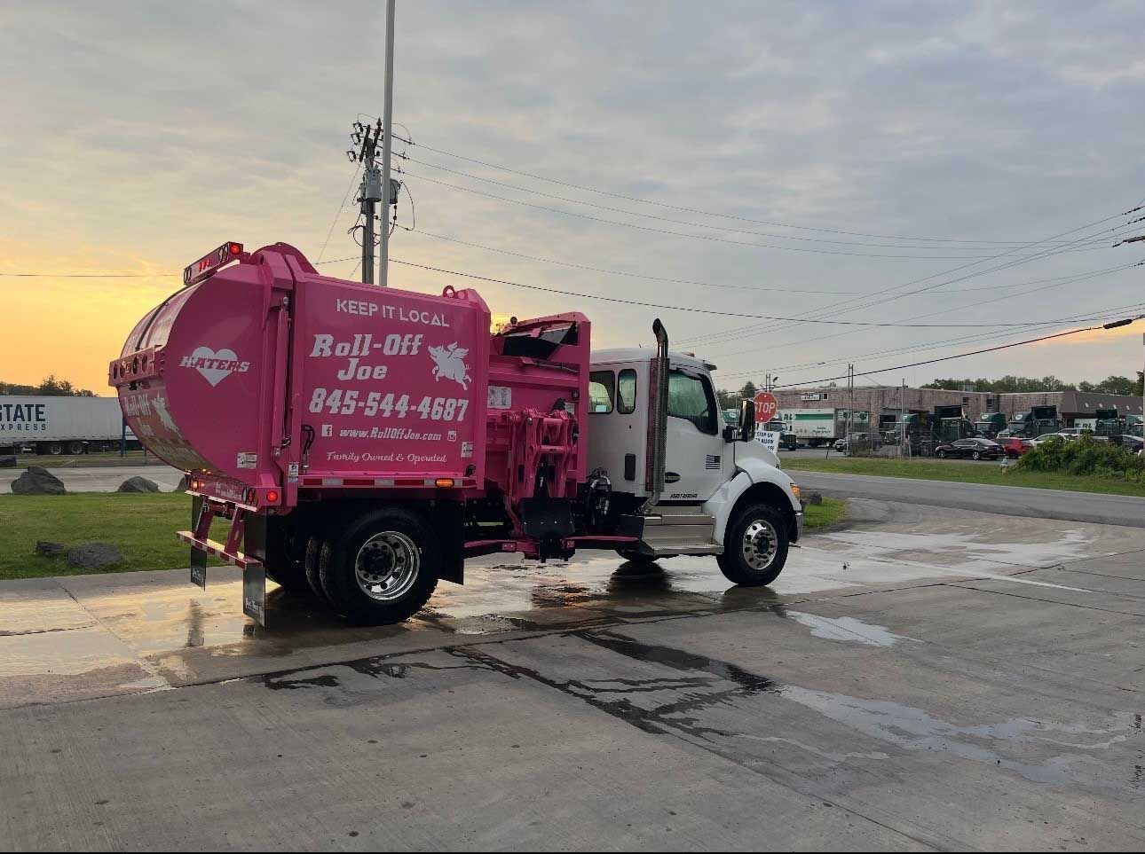 Pink Roll-Off Joe garbage truck, white cab, parked on a wet surface with a cloudy sky backdrop.