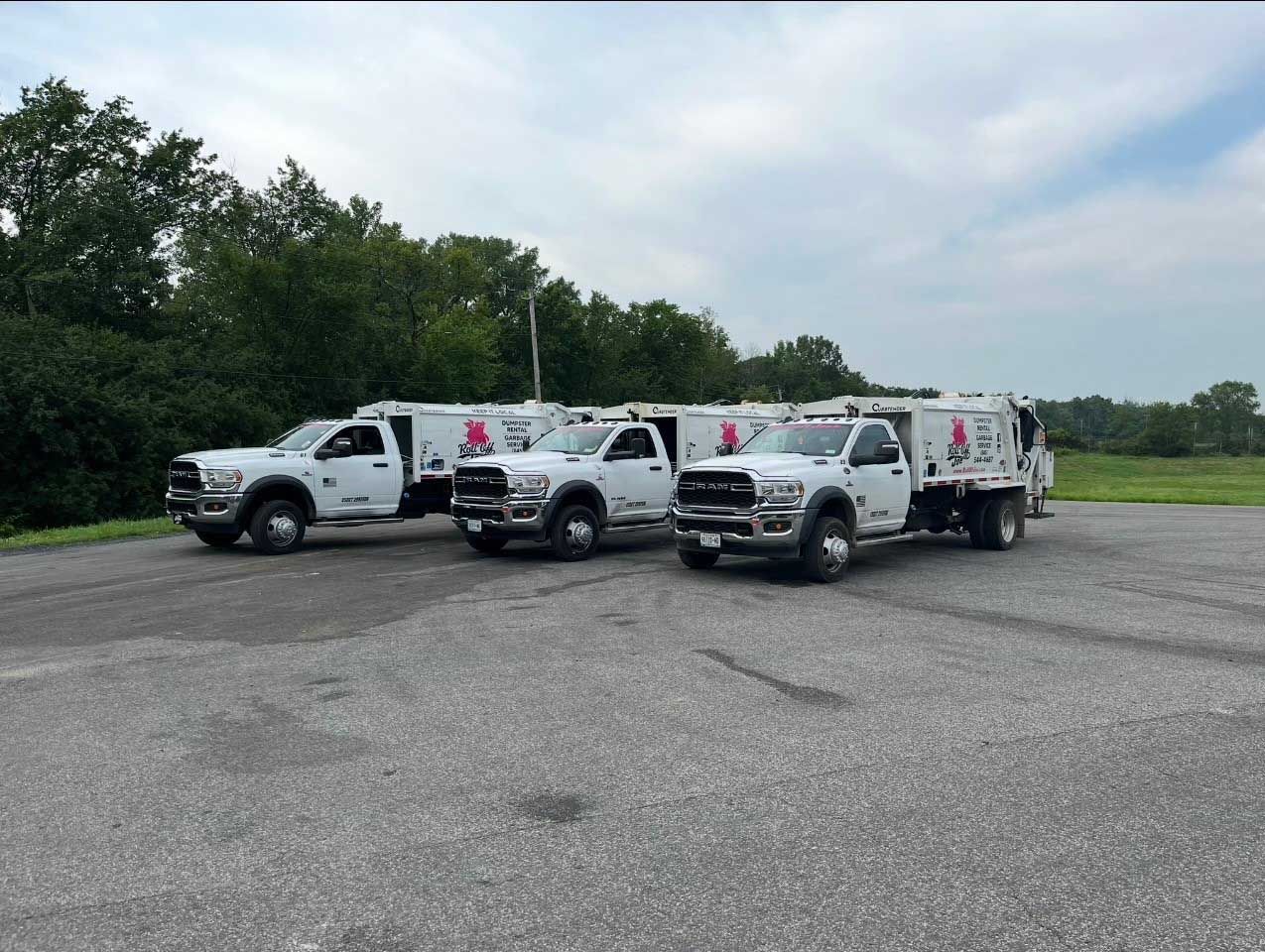 Three white work trucks parked on asphalt, logo visible on doors. Trees and sky in background.