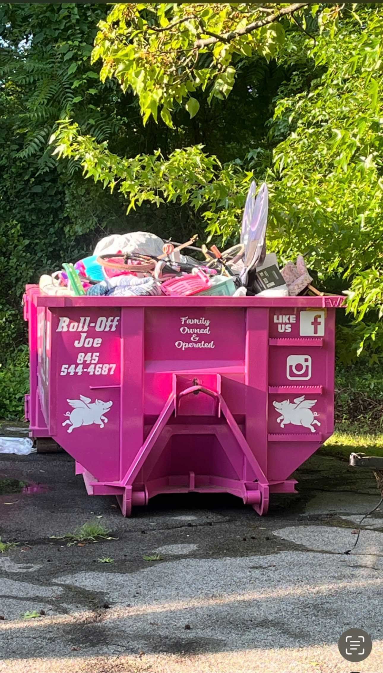 Bright pink dumpster overflowing with trash, in front of a tree.