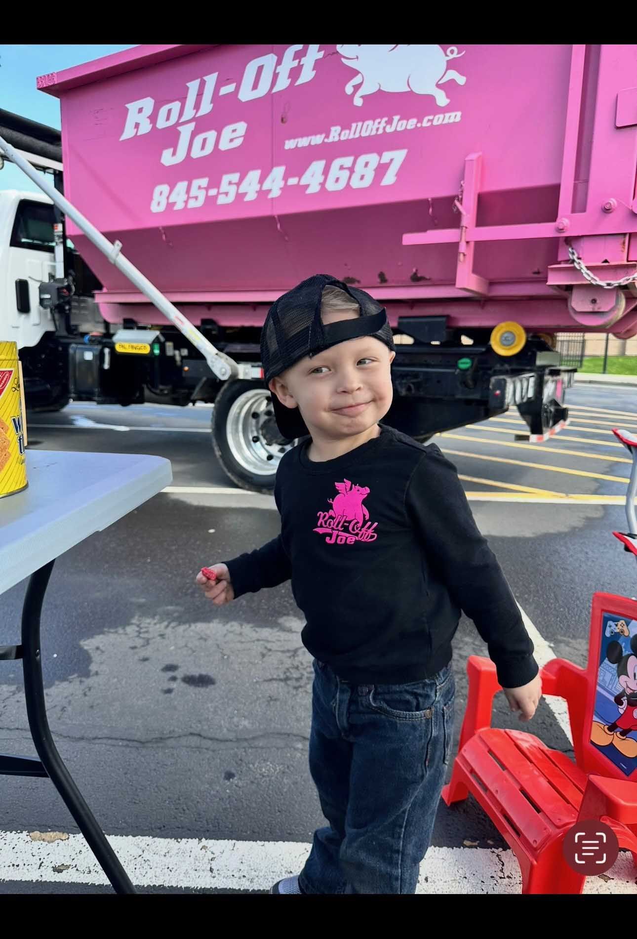 A smiling child wearing a black shirt and jeans stands near a pink dumpster truck.