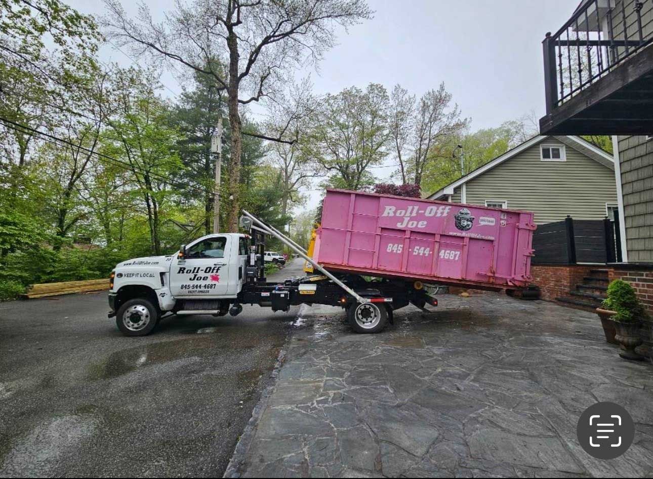 A pink dumpster being lifted by a truck on a driveway, near a house and trees.