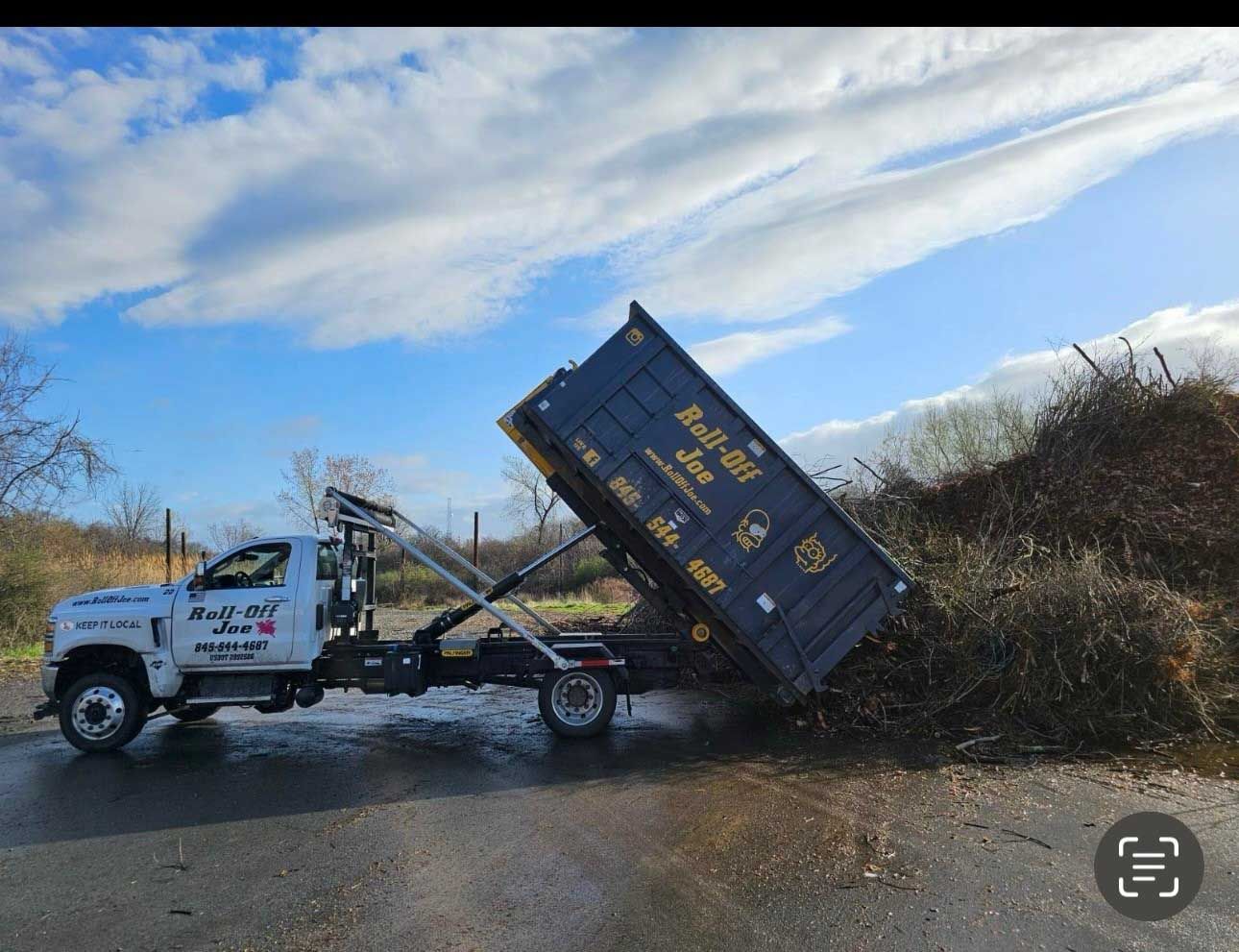 Truck dumping debris from a black container into a large pile outdoors. Cloudy sky in the background.