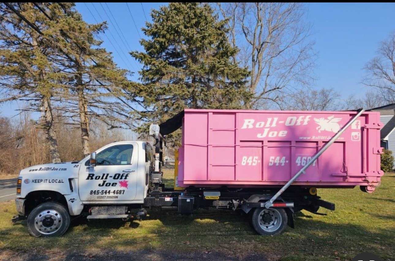 Pink roll-off dumpster on a truck, parked on a grassy area next to a road, under a blue sky.