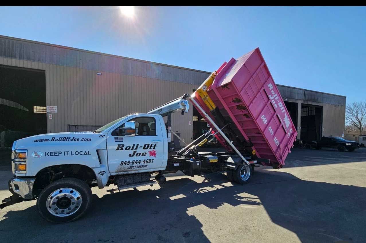 A pink dumpster being lifted by a white roll-off truck, parked in front of a building on a sunny day.