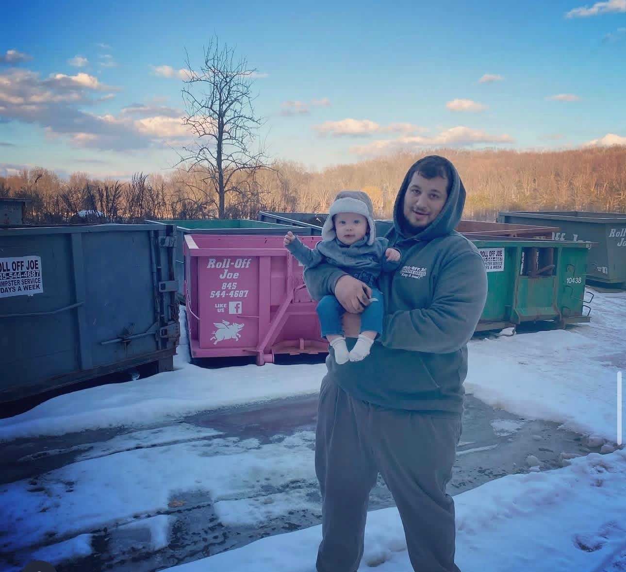 Man holding baby in front of dumpsters, some snow on the ground. Bright sky, outdoor setting.