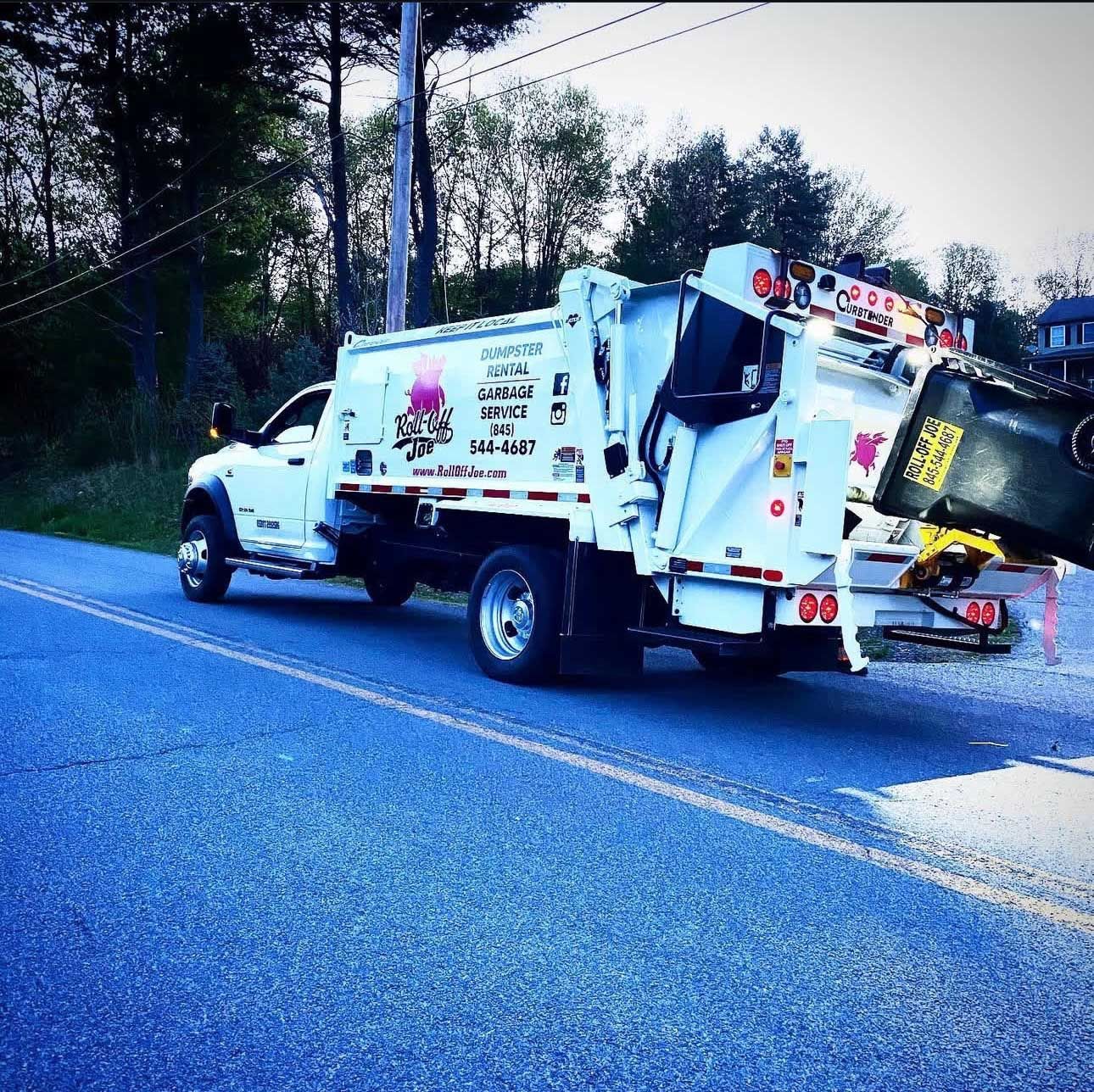 White garbage truck on a road, raising a black trash bin. Pink logo on the truck, trees in the background.