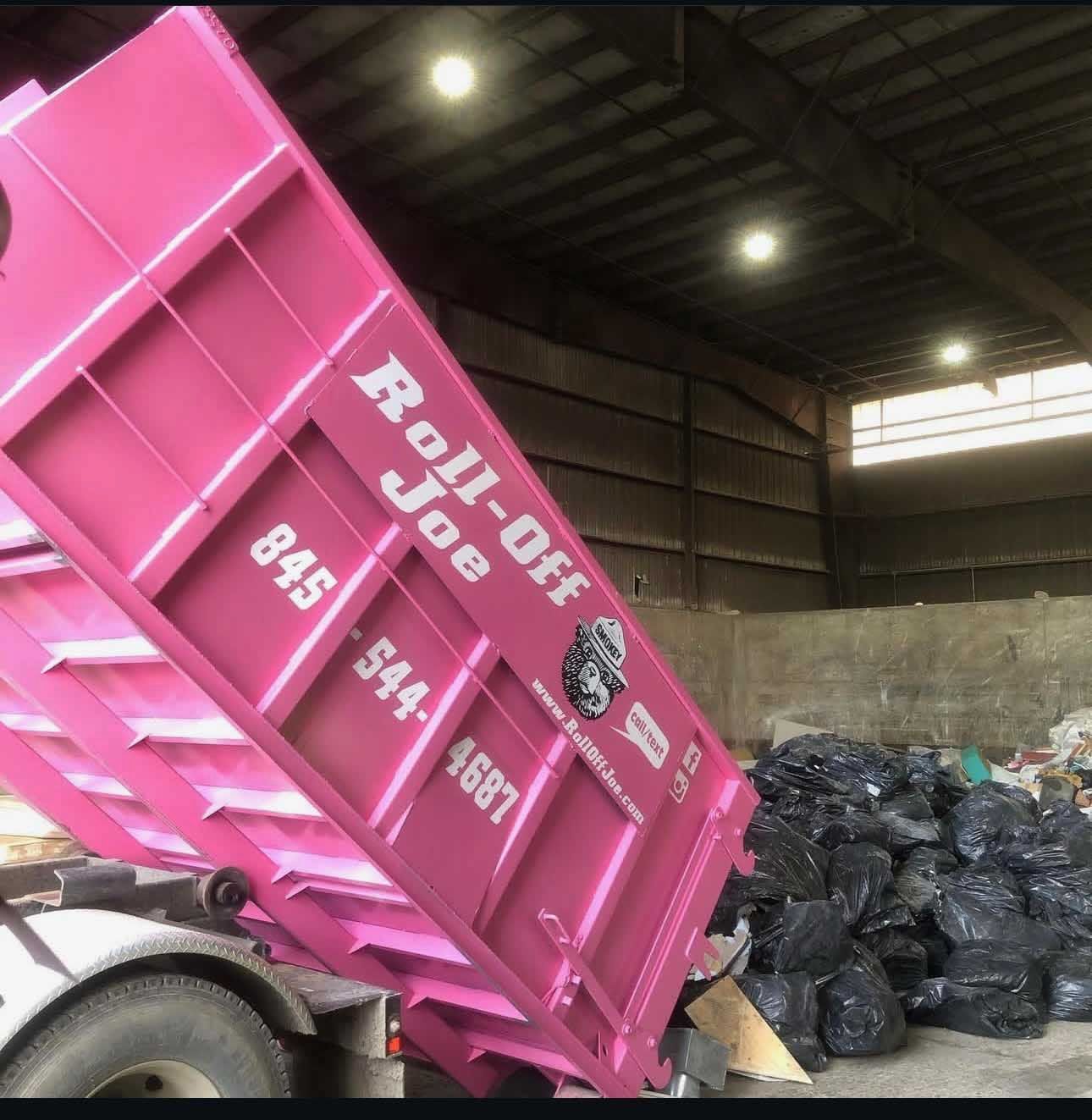 Pink Roll-Off Joe dumpster unloading waste. Dumpster is lifted, revealing bags of trash inside a warehouse.