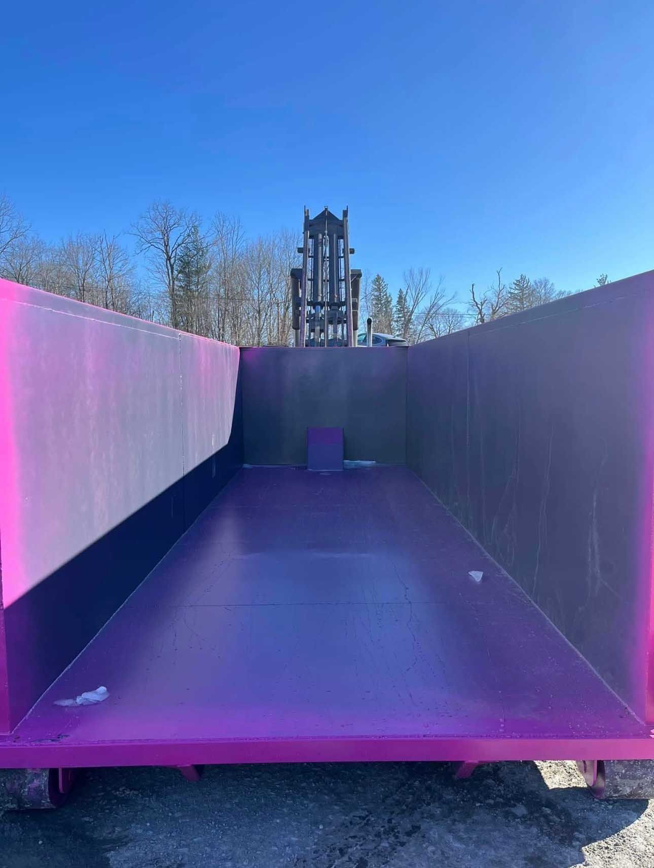 Empty purple dumpster with a dark metal structure in the background under a blue sky.