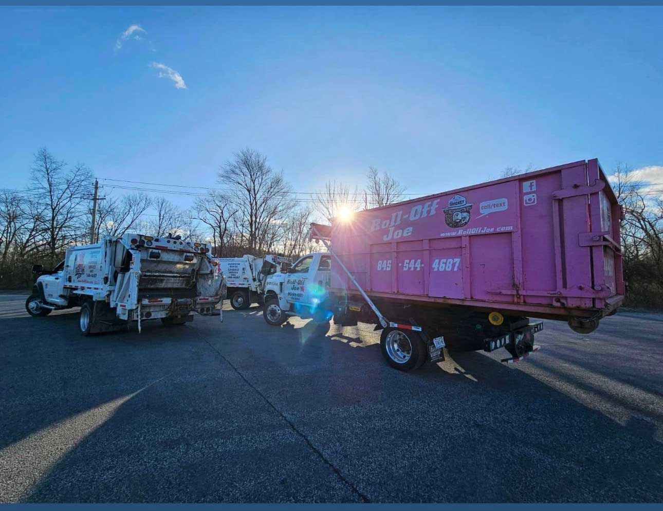 Pink dumpster trailer hitched to a truck, parked with other trucks under a sunny sky.