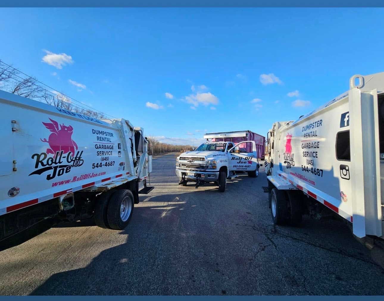 Three garbage trucks and a pickup truck parked on a road with a mountain background.