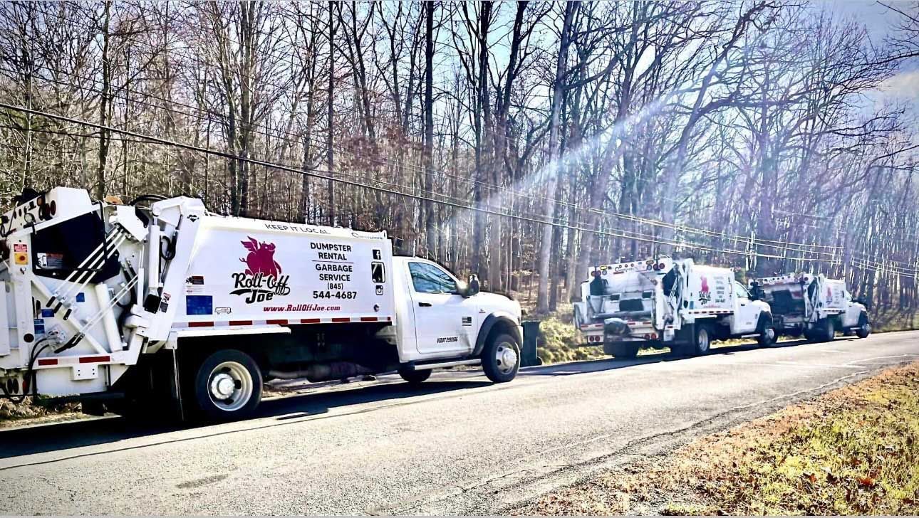 Three white garbage trucks parked on a road lined by leafless trees.