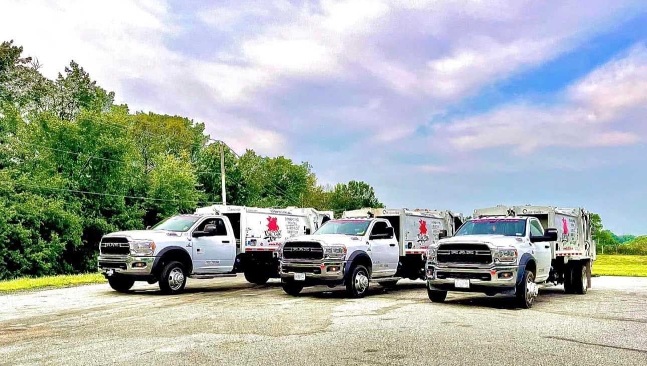 Three white work trucks parked on gravel, with company logo and a cloudy blue sky.