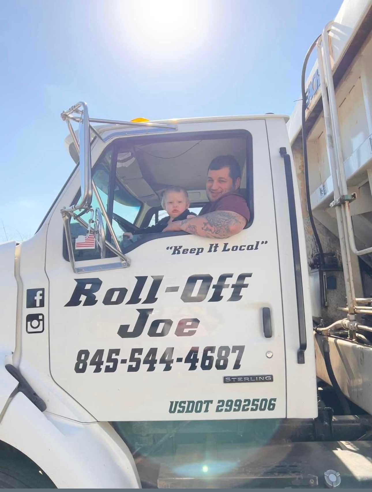 Man and child in a Roll-Off Joe truck cab, under a blue sky. The truck is white with large lettering.