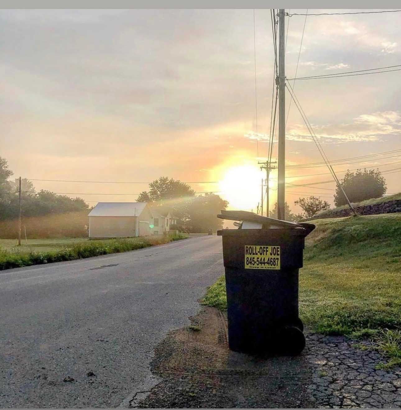 Black trash can on a road, with sunrise in the background. Gray asphalt, green grass, power lines.