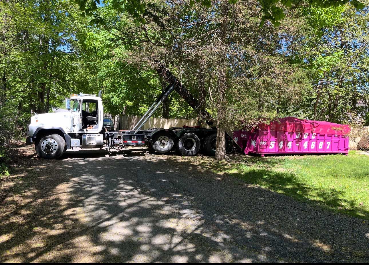 White truck with pink dumpster next to a tree on a gravel driveway.