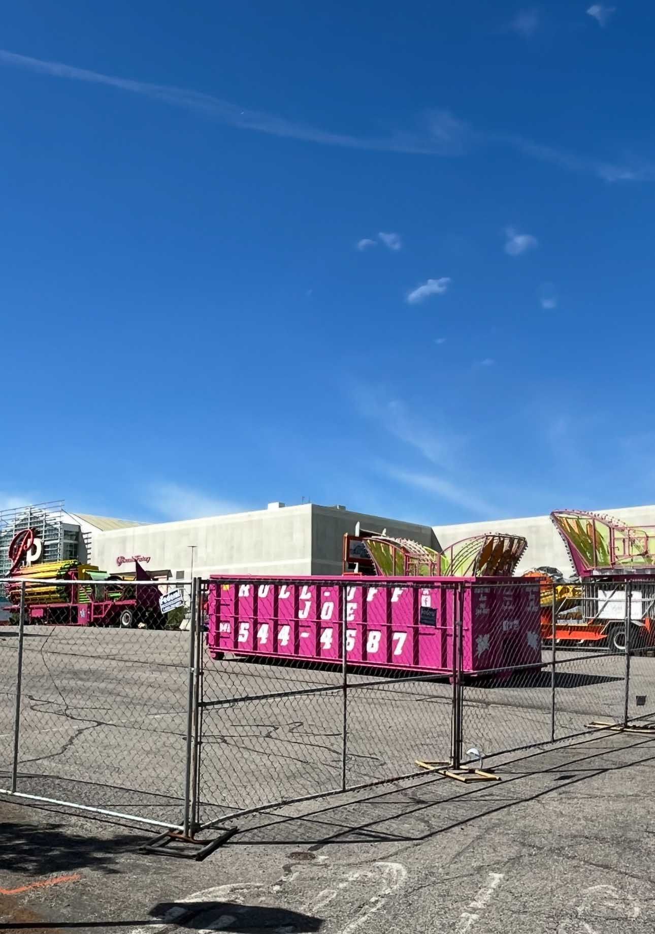 A pink dumpster with numbers sits in a parking lot with amusement park equipment under a blue sky.